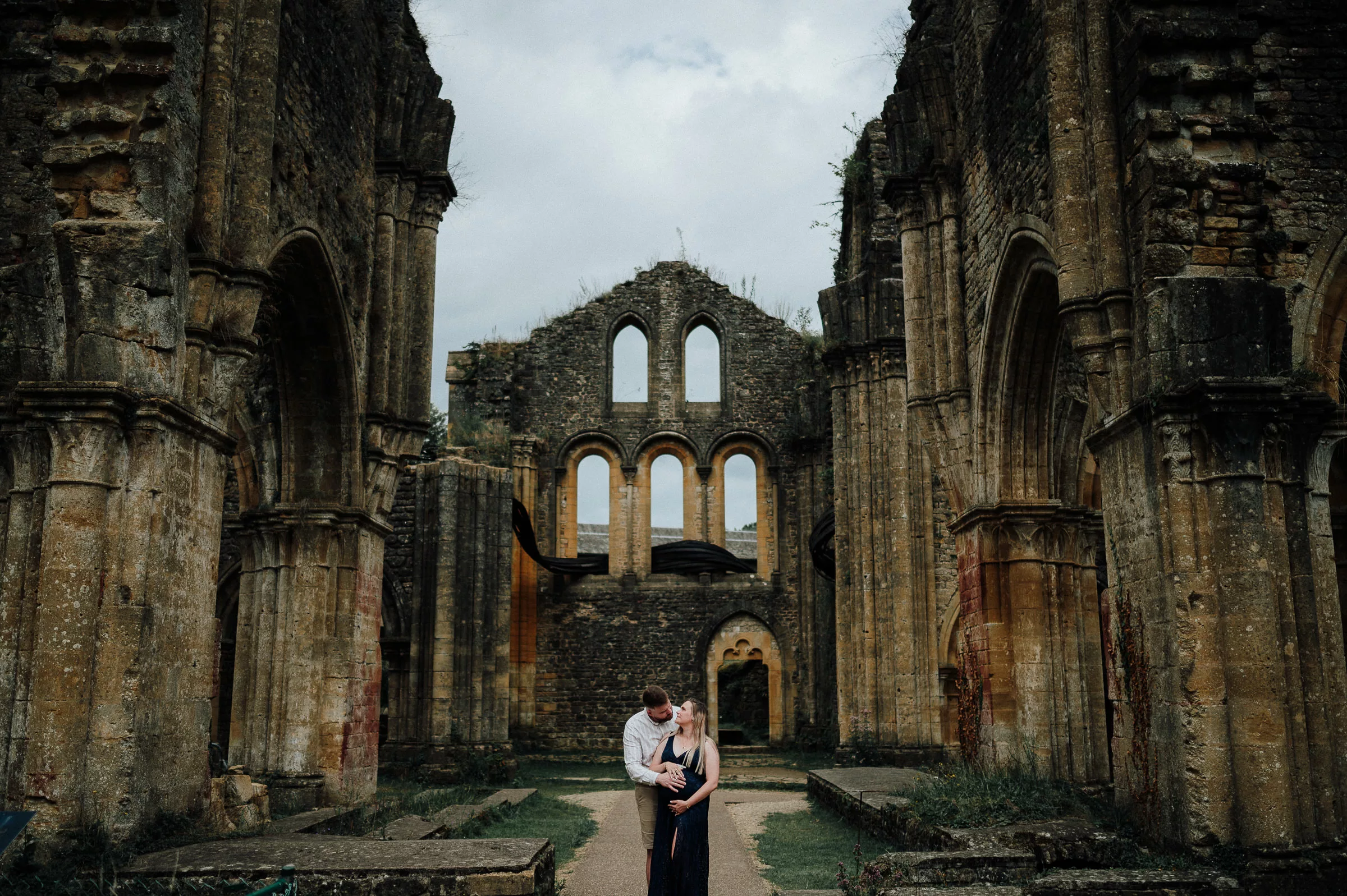 Séance grossesse à l’Abbaye d’Orval – Laurine et Emmanuel dans les ruines de l’abbatiale, lumière douce sur les pierres anciennes